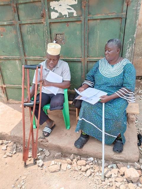 Two community members with mobility aids review programme documents outside a home.