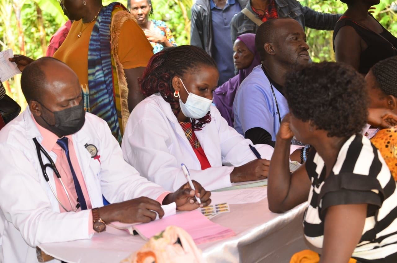 Two clinicians in white coats consult with community members at an RDAF outreach table.