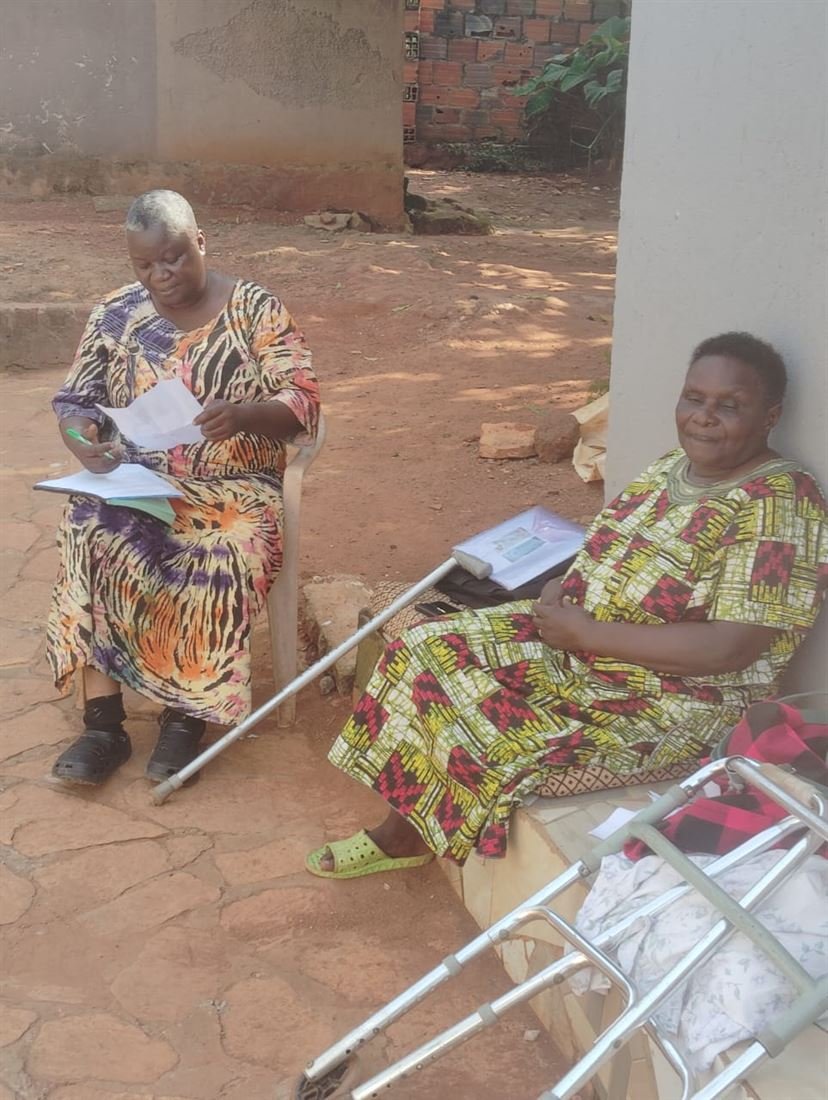 A woman with a walking frame sits beside another community member during an RDAF home visit.
