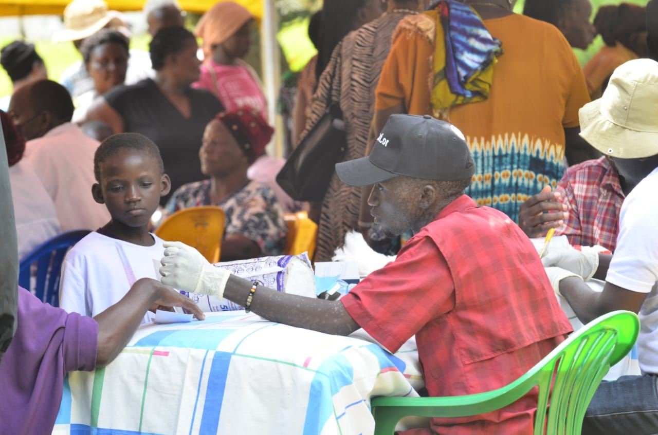 A health worker in gloves performs a finger-prick blood glucose test on a community member's hand at a screening outreach.