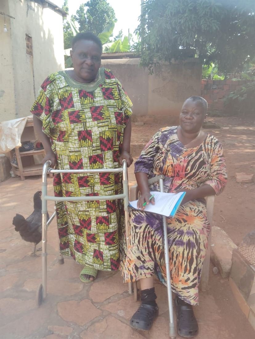 A woman stands supported by a walking frame outside her home while an RDAF team member sits with her.